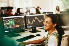 Donna sorridente seduta dietro la sua scrivania, con schermi che visualizzano informazioni finanziarie.