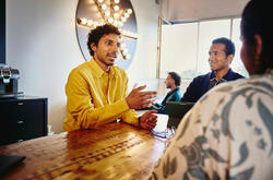 un ragazzo in piedi appoggiato ad un tavolo che parla con un uomo e una donna