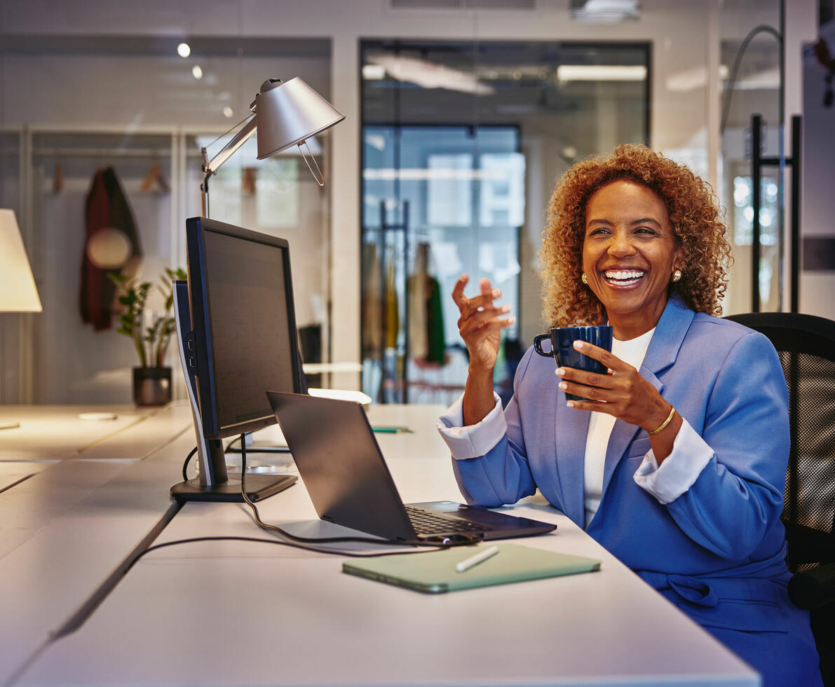 una donna sorridente che beve un caffe alla sua scrivania davanti al pc