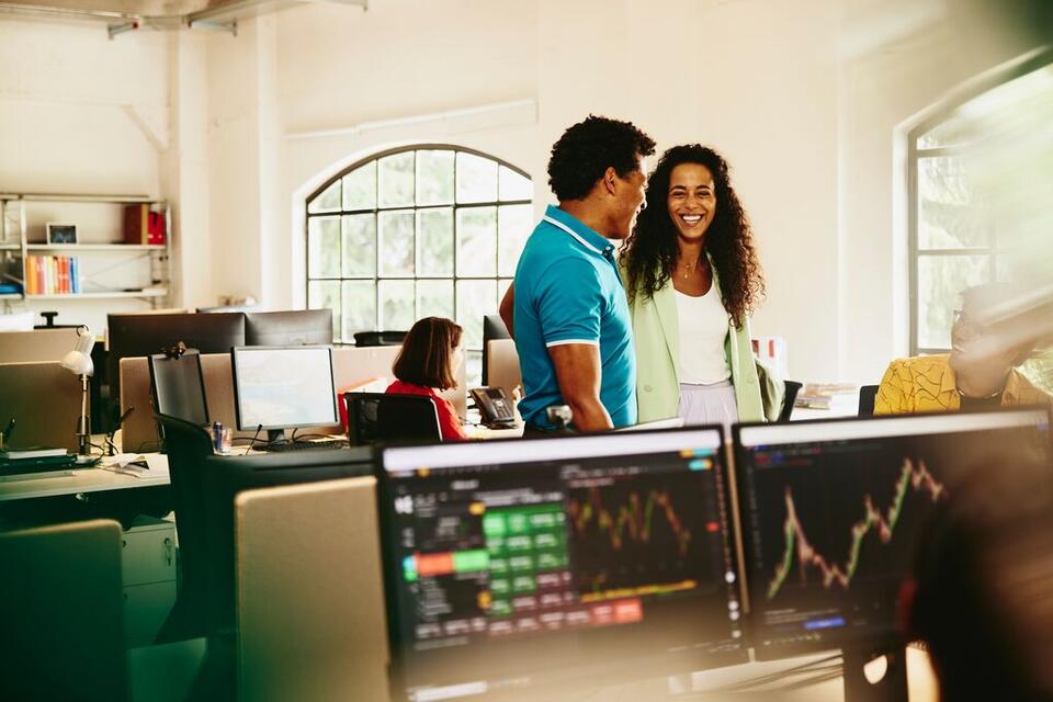 due colleghi che parlano al centro di un ufficio dietro ad alcuni pc con grafici - market intelligence