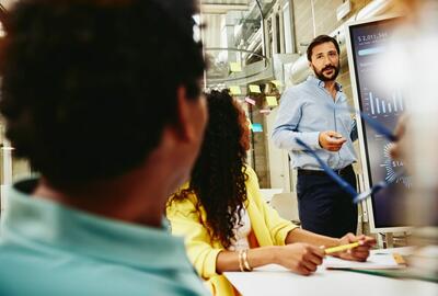 resilienza aziendale: un uomo che tiene una riunione parlando vicino ad una lavagna con grafici, mentre i colleghi lo ascoltano