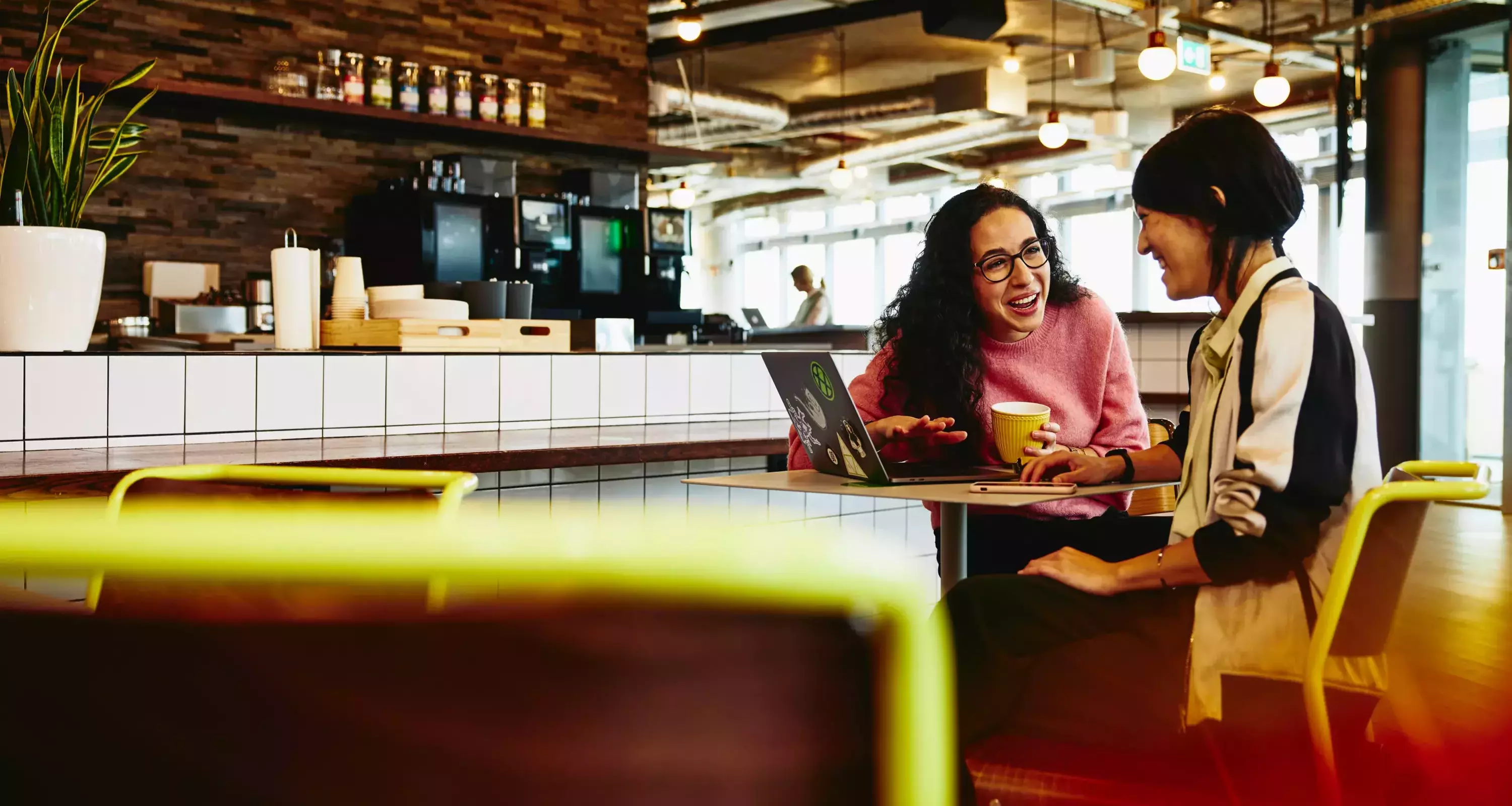 Two female sitting in restaurant having a converstation, looking at laptop, smiling.
